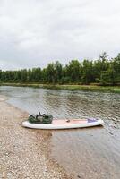 A serene scene with a paddle board resting by a calm riverbank under cloudy skies, perfectly harmonizing with its surroundings to convey a sense of peace and connection with nature photo