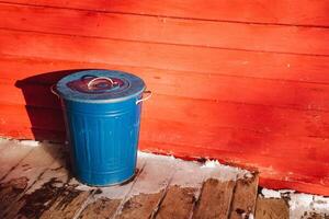 In front of a vibrant red wall, a blue trash can is positioned on a polished wooden floor, showcasing the contrast between the colors and materials in this rooms design photo