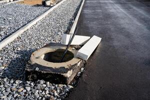 A round manhole cover is positioned on the side of a busy road, just beside the asphalt, where automotive tires frequently pass over it, surrounded by the landscape of soil and concrete photo