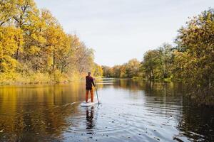 A man is skillfully maneuvering a paddle board on a picturesque river, fully immersing himself in the serene surroundings while appreciating the stunning beauty of nature all around him photo