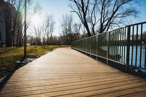 There is a beautiful wooden bridge that gracefully leads to a serene lake nestled within a picturesque park, surrounded by vibrant natural scenery and elements of nature photo