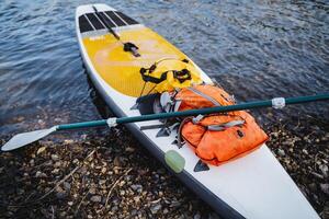 A close-up of a SUP board is moored to a sandy shore, things are collected in a dry bag, a paddle for paddling lies on a sap, a hike along the river. photo