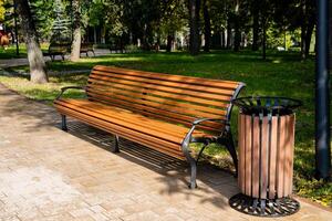 In a serene park setting, there is a sturdy wooden bench complemented by a trash can, providing a place for relaxation and waste disposal amid the natural beauty of the surroundings photo