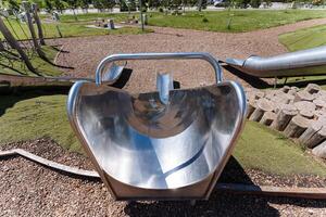 A stainless steel slide is featured at the playground in the park, surrounded by green grass and other elements like plants and trees. Safety equipment like helmets is encouraged for use photo