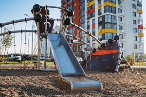 There is a colorful slide in a childrens playground, and in the background, you can see a tall building against the sky accompanied by greenery and a distant view of the city photo