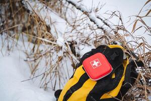 A bright red first aid kit is resting on top of a large yellow backpack that is situated in the soft, white snow all around it, creating a striking contrast against the winter landscape photo