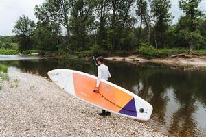 Colorful bright beautiful SUP board in the hands of a man, man preparing to sail down the river on a paddle board, pebble beach on a mountain river. photo