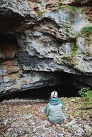 A person is comfortably sitting on the ground in front of a large, mysterious cave, surrounded by various natural elements and an intriguing landscape that captures the imagination photo