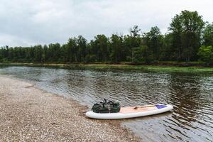 A beautiful landscape on the river surrounded by forest, a SUP board stands alone on the river bank, an inflatable board with an oar. photo