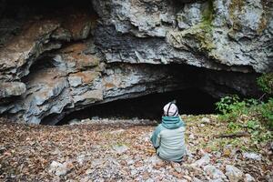 A person is comfortably seated on the ground in front of a large cave entrance, enjoying the natural beauty of the surroundings and the unique geological features nearby photo