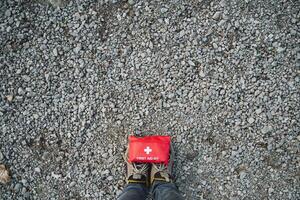An Emergency First Aid Kit is placed on a gravel surface, paired with sturdy hiking boots, ideal for outdoor exploration and prevention photo