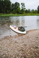 A lonely SUP stands on the bank of a mountain river against the background of a forest, a white inflatable board with an oar. photo