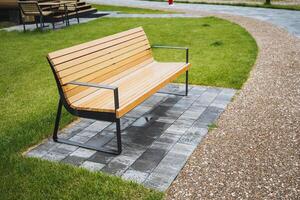 In a peaceful park, a basic wooden bench sits on a red brick path amidst green grass and a stunning natural backdrop, providing a serene spot to relax and admire the surroundings photo