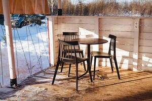 A beautifully crafted wooden table, accompanied by its matching chairs, is elegantly arranged on a serene patio blanketed in fresh, white snow, forming a peaceful and inviting winter scene photo
