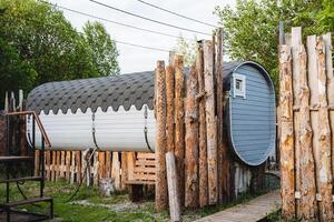 A barrelshaped structure is nestled within the scenery, enclosed by a wooden fence, effortlessly fitting in with the lush landscape featuring trees, grass, and a house topped with a roof photo