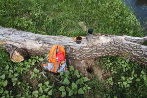 Camping equipment top view, hiking in the forest, backpack lying on the grass, mug with thermos standing on a log. photo