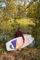 A man holds a SUP board in his hand and goes down to the river, SUP surfing on a reservoir, a solo trip across the lake on a paddle board. photo