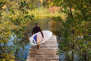 A man is carefully carrying a paddle board while walking on a dock that extends over the water, surrounded by a beautiful natural landscape and lush greenery of trees alongside the lake photo