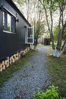 A black house with a white window is located peacefully amidst trees, grass, a gravel path, in a serene natural setting. It showcases a beautiful blend of nature and architecture photo
