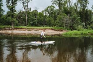 The man finds immense happiness paddle boarding in a tranquil river, surrounded by lush greenery under a blue sky, feeling a deep connection with the natural beauty surrounding him photo