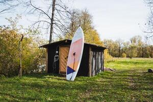 A colorful surfboard leans against a rustic shed, adding to the tranquil scene surrounded by lush green grass, enhancing the peaceful atmosphere of this picturesque setting photo