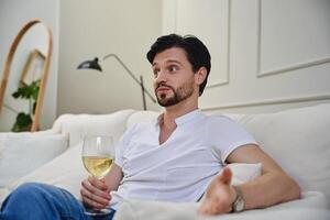Man enjoying a glass of white wine while sitting on a couch at home photo