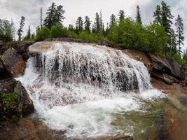 Cascades of a powerful waterfall in a green mountain forest. Nature background of turbulent falling water stream on wet rocks. Taigishonok River, Western Sayans. photo