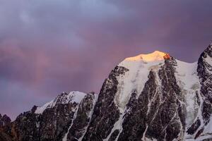 Magnificent purple sunset in the mountains. Red fiery cloud in the evening over the silhouette of sharp mountains. Top black rocks in evening golden sunshine and white-snow pointy peak. photo