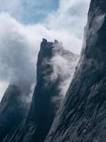 Vertical view of great mountains in the fog. Low clouds and dark peaks of the mountains. Atmospheric surreal landscape with dark rocky mountain top in low clouds in cloudy sky. photo