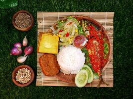 Top view, complete Mixed Rice served with white rice, sambal fish, tempeh, tofu, fresh vegetables, sambal, and spices on a bamboo mat. photo