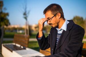 Tired businessman having a headache while sitting on the bench in the park photo