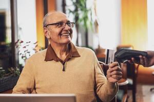 Cheerful senior man with eyeglasses in casual clothing making a contactless payment with credit card while sitting in the cozy city cafe at evening. photo