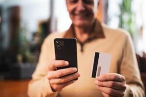 Close up photo of senior man using mobile phone and credit card while sitting in cafe. Focus on credit card.