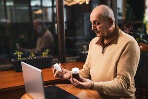 Uncertain senior man having online consultation with his doctor on laptop while showing him two bottle of pills. He is asking for advice for his health problems while sitting in the cafe. photo
