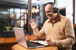 Happy senior man with wireless headphones using wireless headphones, laptop and taking notes while attending online class. He is waving and talking during a call on computer. photo