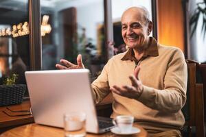 Joyful senior man in casual clothing talking and gesturing while having a call on his laptop. He is using computer and enjoying drinking coffee and relaxing in the city cafe. photo