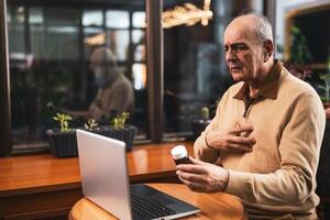Worried senior man holding bottle of pills,using laptop and talking with his doctor during call. He is sitting in cafe and explaining his medical condition while taking this medicine. photo