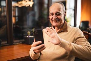 Cheerful senior man in casual clothing waving while using mobile phone and earphones. He is relaxing in the cozy city bar and using technology. photo