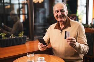 Smiling senior man in casual clothing using mobile phone and credit card while enjoying drinking coffee in the cozy city cafe. photo