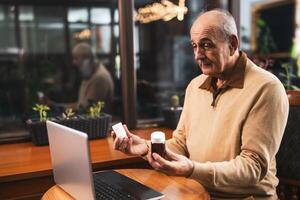 Confused senior man having online consultation with his doctor on laptop while showing him two bottle of pills. He is asking for advice for his health problems while sitting in cafe. photo