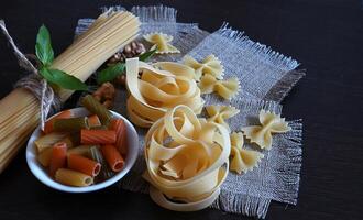 A still life image showing spaghetti, a red bow, ripe tomatoes, a block of cheese, garlic, and fresh basil all laid out on a dark wooden surface, with additional dry pasta and seasonings. photo