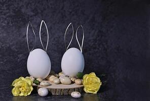 This image features two white eggs with paper bunny ears, arranged on a rustic wooden slice alongside small speckled stones and yellow flowers. photo