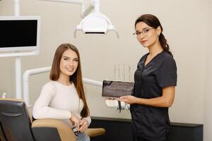 Woman dentist demonstrating tablet with treatment plan to worried female patient in chair clinic. Dentistry visitor looking on gadget screen photo