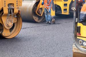 A road worker corrects freshly rolled asphalt with a shovel against the backdrop of the work of road rollers. photo