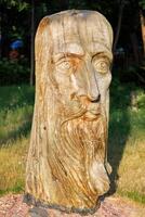 Wooden sculpture of the head of an elderly man lit by warm sunlight against a blurred background of a green park. 06.19.2022. Kyiv. Ukraine. photo