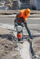 A builder in an orange cape is ramming a trench with a vibratory rammer at a construction site. photo