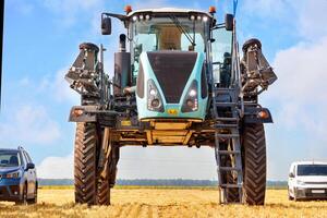 A tractor sprayer with a high wheelbase stands next to cars in a field against a blue cloudy sky. photo