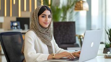 Portrait of a Stylish and Confident Young Muslim Female Manager Sitting at her Desk and Using a Laptop Computer in a Modern and Professional Office Environment photo