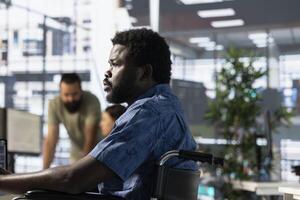 Person with paraplegia sitting in wheelchair at desk in office, doing research for team project. Man with disability looking at computer screen in inclusive workplace, solving tasks photo