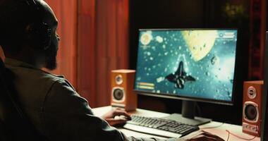 African american man wearing headphones playing SF space videogame on PC screen in blurry background, shooting lasers. Close up of person enjoying galaxy flying game on computer monitor, camera B photo